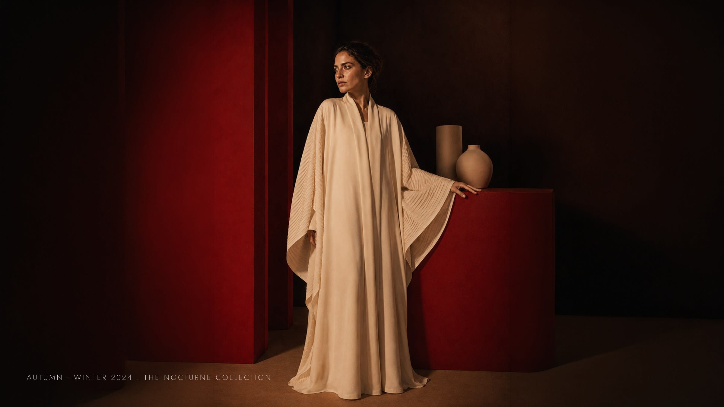 A cream pleated abaya beside a red plinth and pale vases.