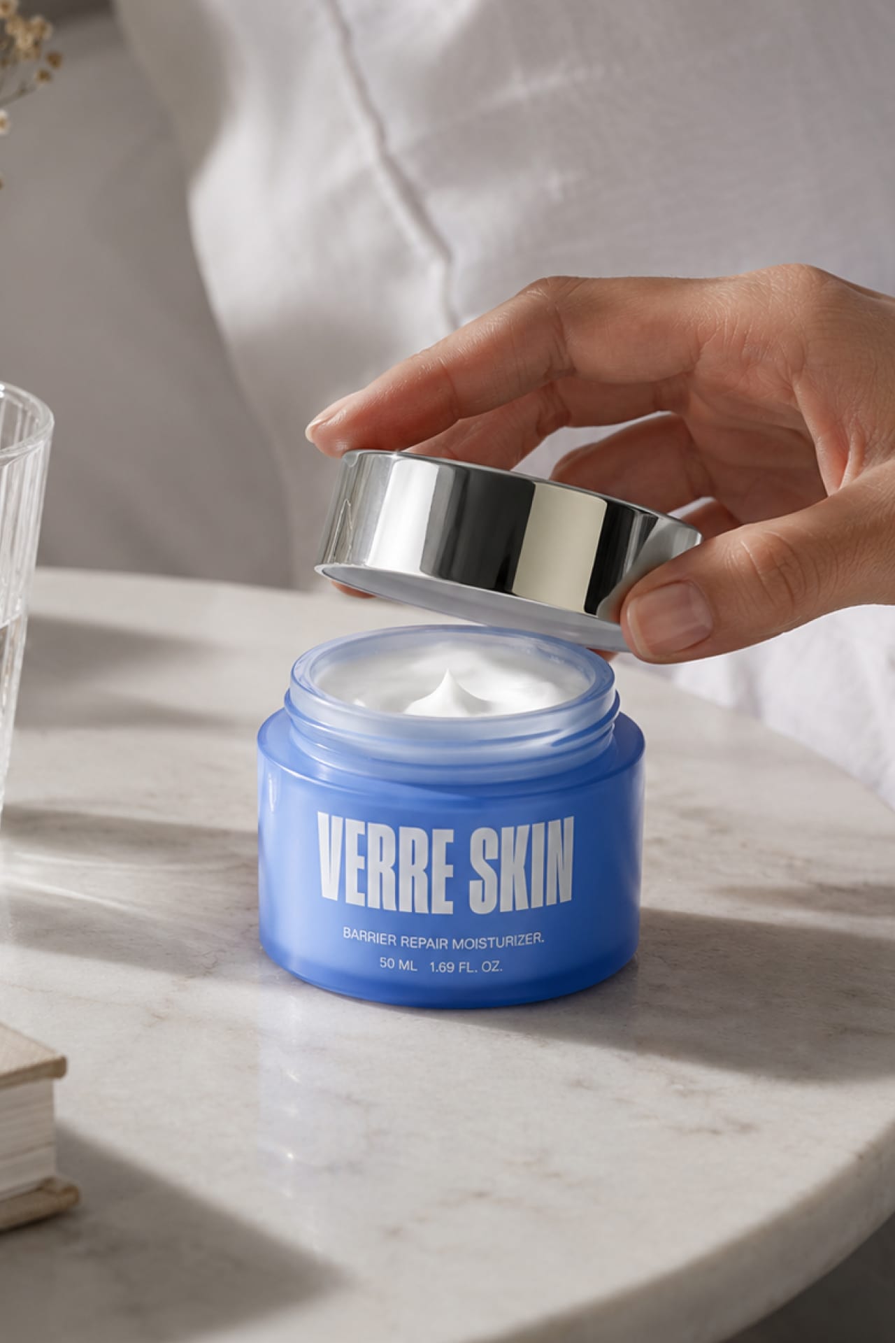 A hand lifting the silver lid off the moisturizer jar of white cream.