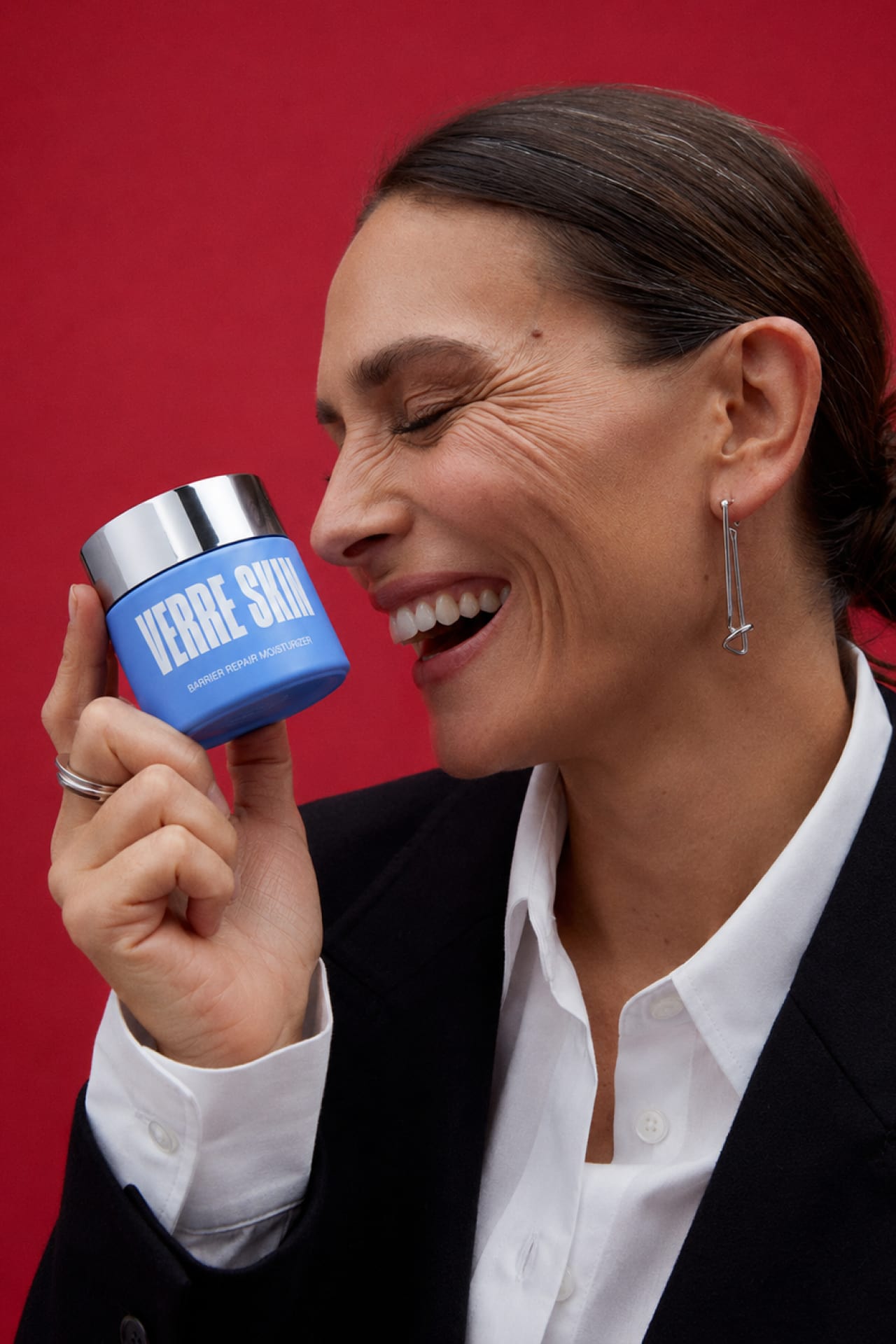 A laughing woman holding the moisturizer jar against a red backdrop.